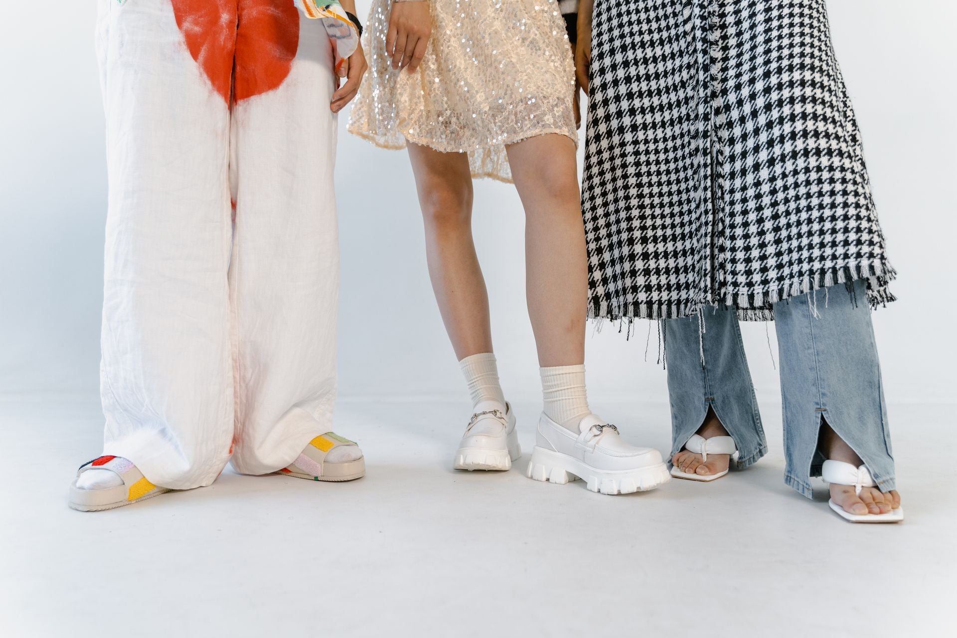 Three models standing together, showing their lower bodies and footwear. The left model wears white wide-leg pants with a bright orange tie and colorful sneakers.
